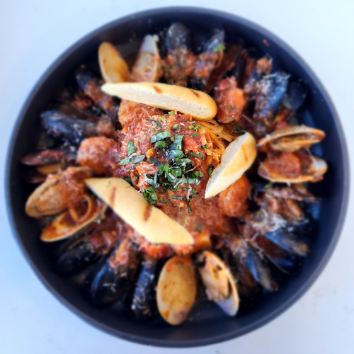 The image shows a bowl of seafood pasta, featuring mussels, clams, a tomato sauce, herbs, and slices of bread on top, all in a black bowl.